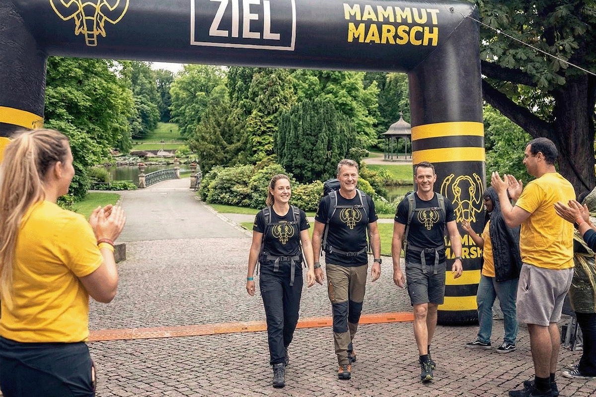 Glückliche Teilnehmer beim Mammutmarsch Zieleinlauf unter dem großen schwarzen Zielbogen im Park nach ihrer Langstreckenwanderung.